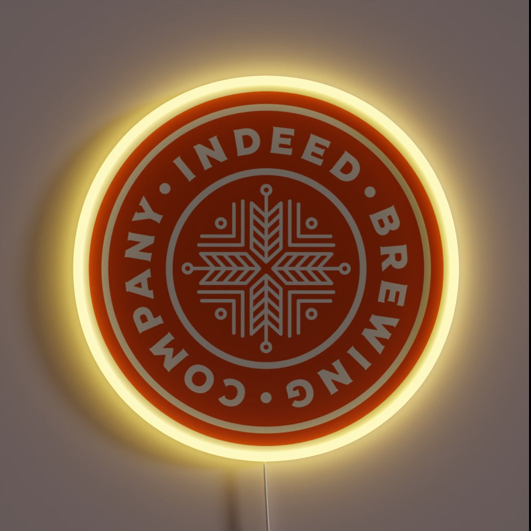 indeed-brewing-company-yellowpop-93558 – Round neon sign with an orange background and white snowflake-style logo for Indeed Brewing Company, outlined in soft yellow glow